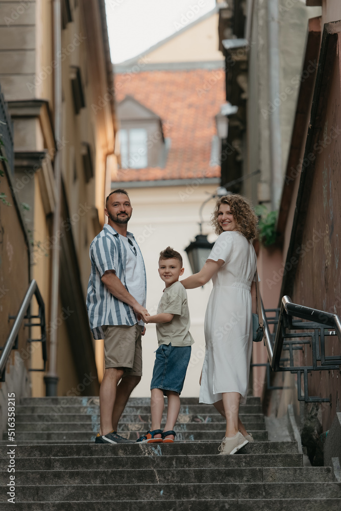 Obraz premium A full-length photo from the back of a family which is turning around while climbing stairs in an old European town. A happy father, mother, and son are holding hands and having fun in the evening.