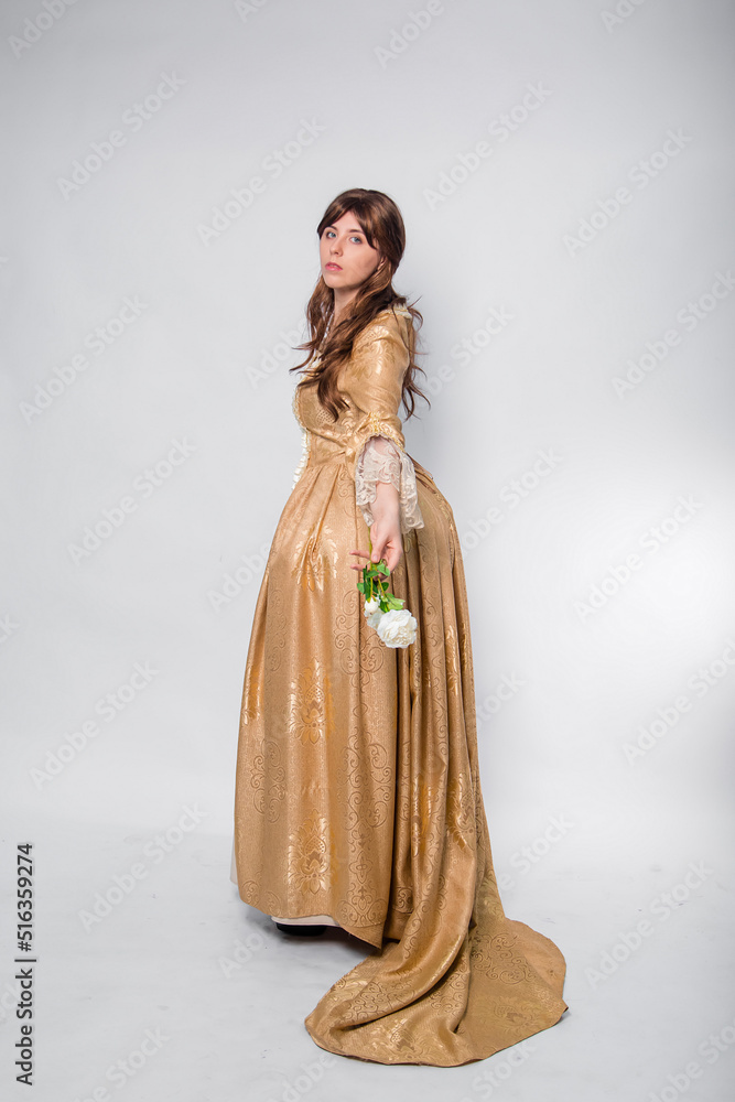 Full length portrait of a woman in a gold dress in the style of the rococo era, standing with her back forward and posing with a flower in hand isolated on a white background.
