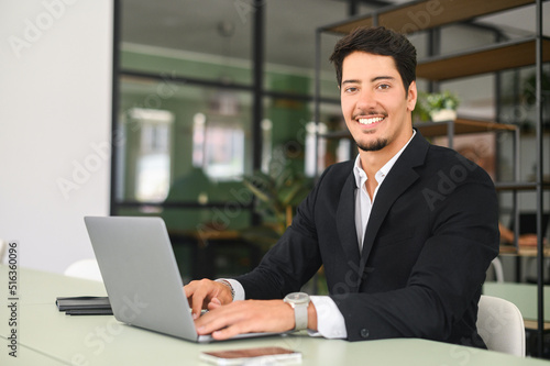 Confident latin businessman using laptop in the office. Young male office employee in formal wear typing and chatting online, develops software indoors, looks at camera with smile