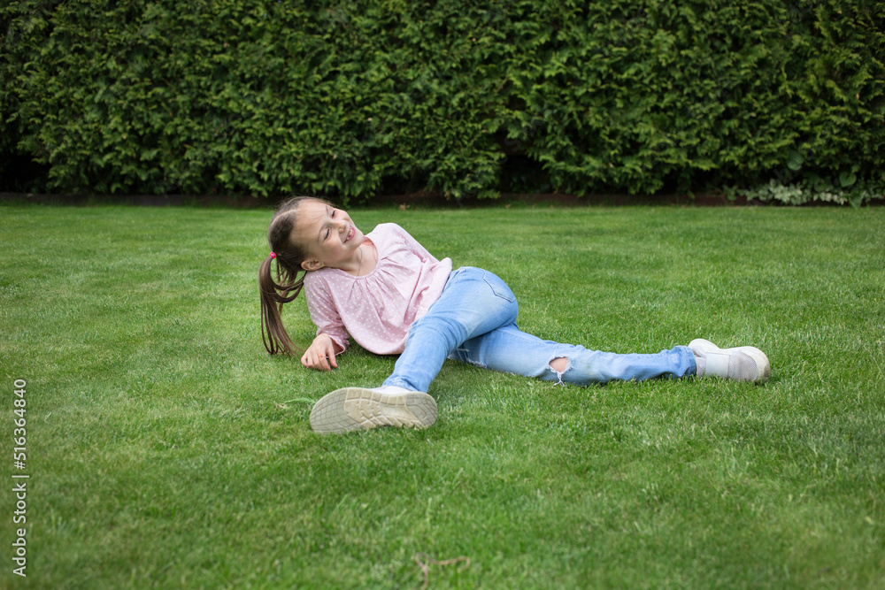 Happy pretty girl 6-7 years old in jeans lies on the lawn in the city park. green nature background. Childhood outdoors, summer, spring pastime in nature Happy pretty girl 6-7 years old in jeans lies on the lawn in the city park. green nature background. Childhood outdoors, summer, spring pastime in nature