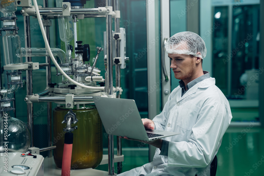 Scientist holding laptop and controlling rotational vaporizer during CBD oil extraction, hemp oil extraction process. Scientist holding laptop and controlling rotational vaporizer during CBD oil extraction, hemp oil extraction process.