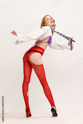 Portrait of young girl in white shirt, tie and red tights posing isolated over grey studio background. Femininity