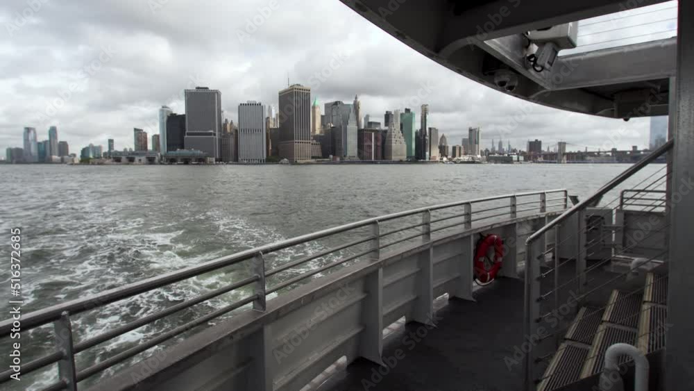 View from NYC ferry, sailing from Manhattan. Foamy trail behind. View of the Manhattan island and famous high massive skyscrapers. Cloudy summer day. USA View from NYC ferry, sailing from Manhattan. Foamy trail behind. View of the Manhattan island and famous high massive skyscrapers. Cloudy summer day. USA