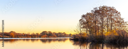 Autumn landscape with trees by the river in sunny weather in warm autumn tones. Reflection of trees in the river