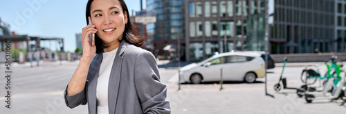 Young happy Asian successful businesswoman entrepreneur wearing suit standing in big city talking on mobile phone. Smiling woman making business call on cell walking on busy street outside.