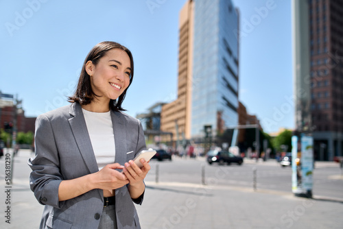 Young smiling professional Asian business woman entrepreneur wearing suit holding smartphone looking at mobile phone using apps on cellphone tech ordering taxi standing on urban city street.