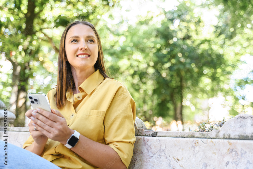 Serene young woman using mobile phone sitting in the park, carefree attractive lady in yellow casual shirt enjoys chatting online, using mobile app Serene young woman using mobile phone sitting in the park, carefree attractive lady in yellow casual shirt enjoys chatting online, using mobile app