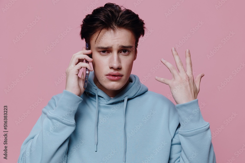a handsome, thoughtful, puzzled man stands on a pink background in a light blue hoodie and holding a smartphone in his hands talks on it, actively gesticulating with his hand. a handsome, thoughtful, puzzled man stands on a pink background in a light blue hoodie and holding a smartphone in his hands talks on it, actively gesticulating with his hand.