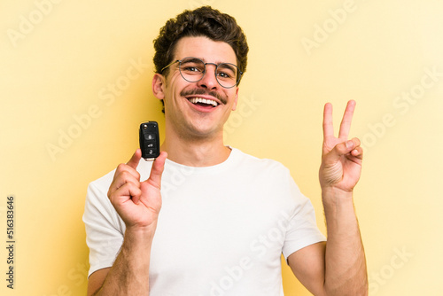 Young caucasian man holding car keys isolated on yellow background joyful and carefree showing a peace symbol with fingers.