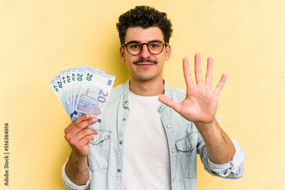 Young caucasian man holding banknotes isolated on yellow background smiling cheerful showing number five with fingers. Young caucasian man holding banknotes isolated on yellow background smiling cheerful showing number five with fingers.