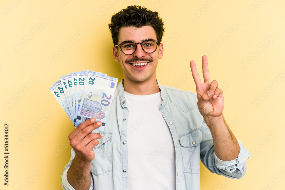 Young caucasian man holding banknotes isolated on yellow background joyful and carefree showing a peace symbol with fingers. Young caucasian man holding banknotes isolated on yellow background joyful and carefree showing a peace symbol with fingers.