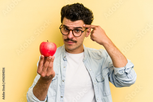 Young caucasian man holding an apple isolated on yellow background pointing temple with finger, thinking, focused on a task. photo