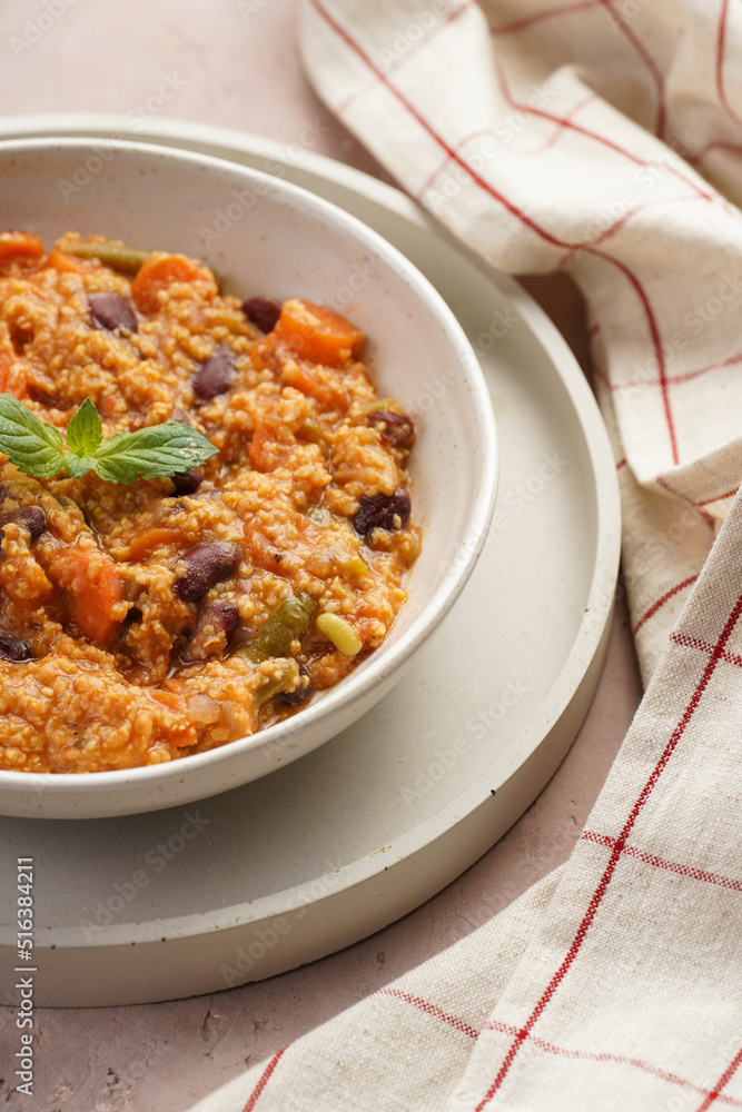 Grain dish - Millet with tomatoes, beans, carrots and spices, mint in a white bowl on a concrete round tray with checkered linen kitchen towel on purple background Grain dish - Millet with tomatoes, beans, carrots and spices, mint in a white bowl on a concrete round tray with checkered linen kitchen towel on purple background