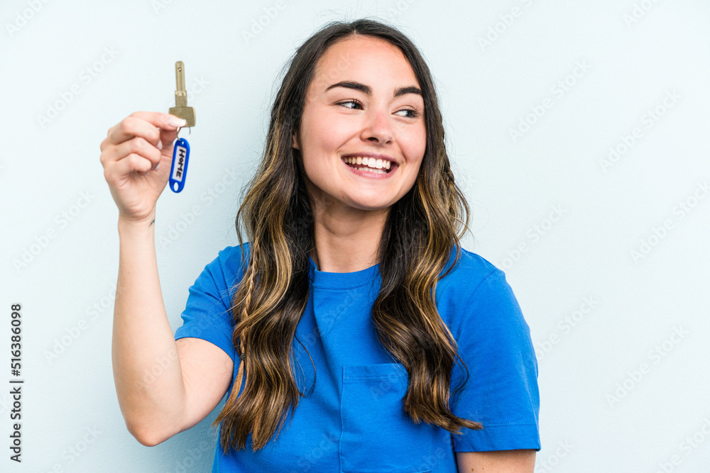 Young caucasian woman holding home keys isolated on blue background looks aside smiling, cheerful and pleasant. Young caucasian woman holding home keys isolated on blue background looks aside smiling, cheerful and pleasant.