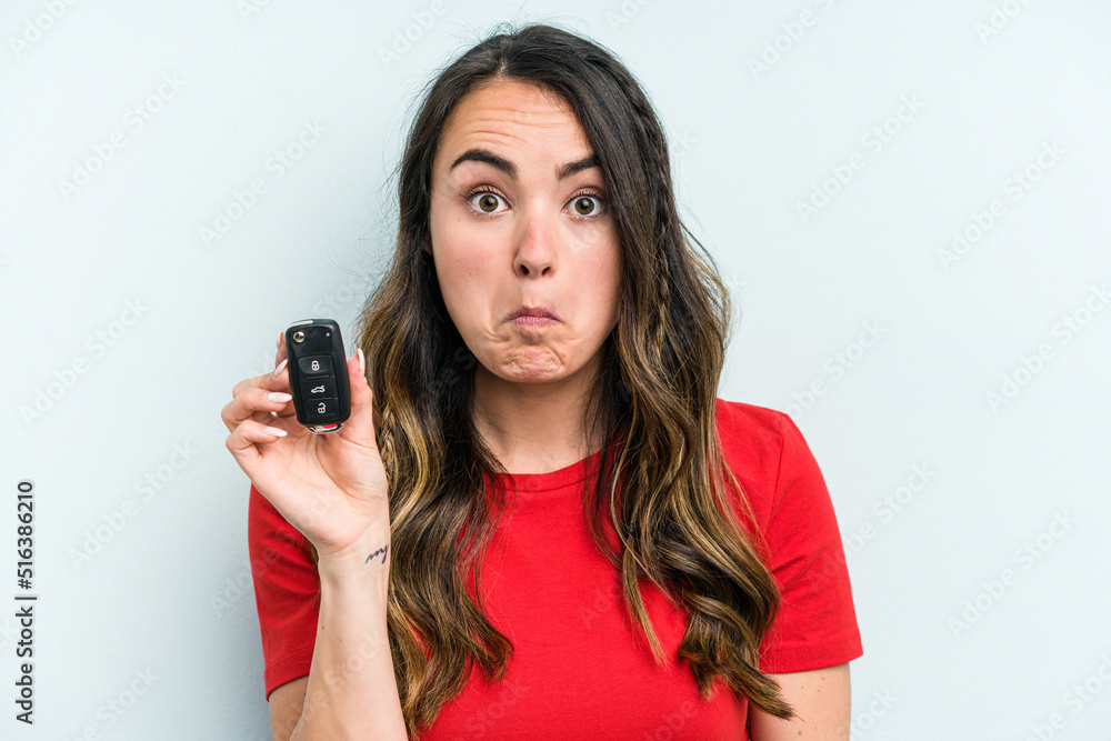 Young caucasian woman holding car keys isolated on blue background shrugs shoulders and open eyes confused.