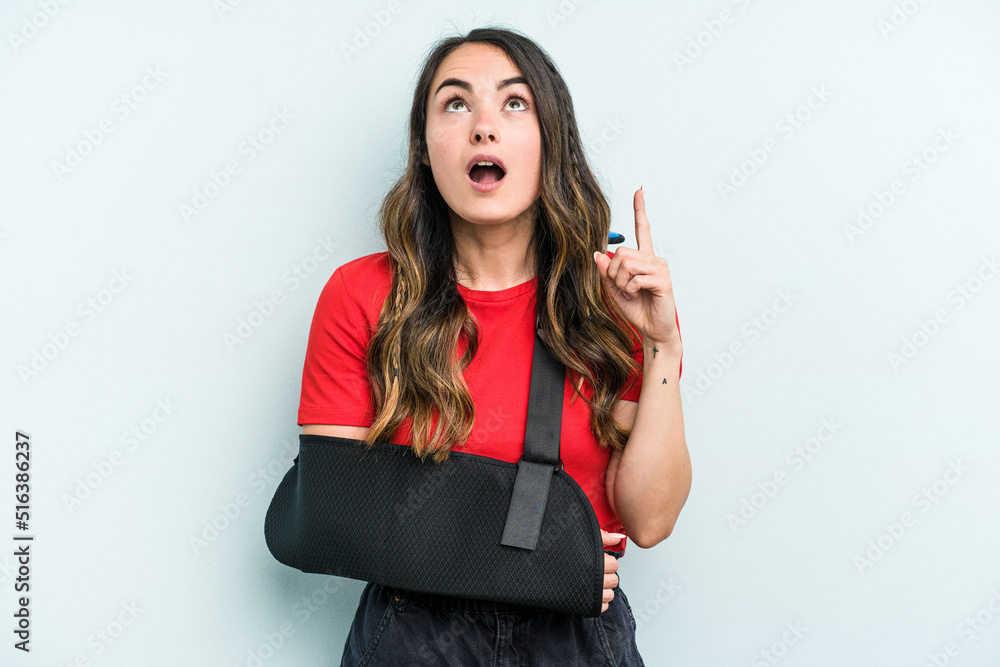 Young caucasian woman with broke hand isolated on blue background pointing upside with opened mouth. Young caucasian woman with broke hand isolated on blue background pointing upside with opened mouth.
