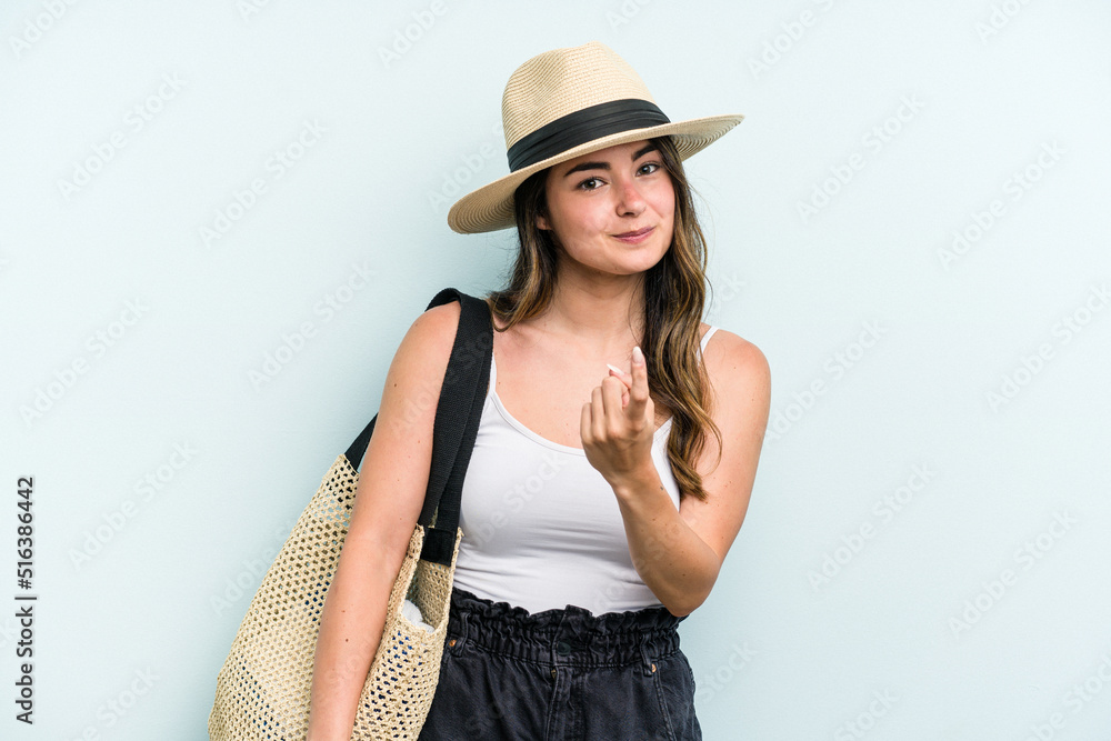 Young caucasian woman holding beach bag isolated on blue background pointing with finger at you as if inviting come closer. Young caucasian woman holding beach bag isolated on blue background pointing with finger at you as if inviting come closer.
