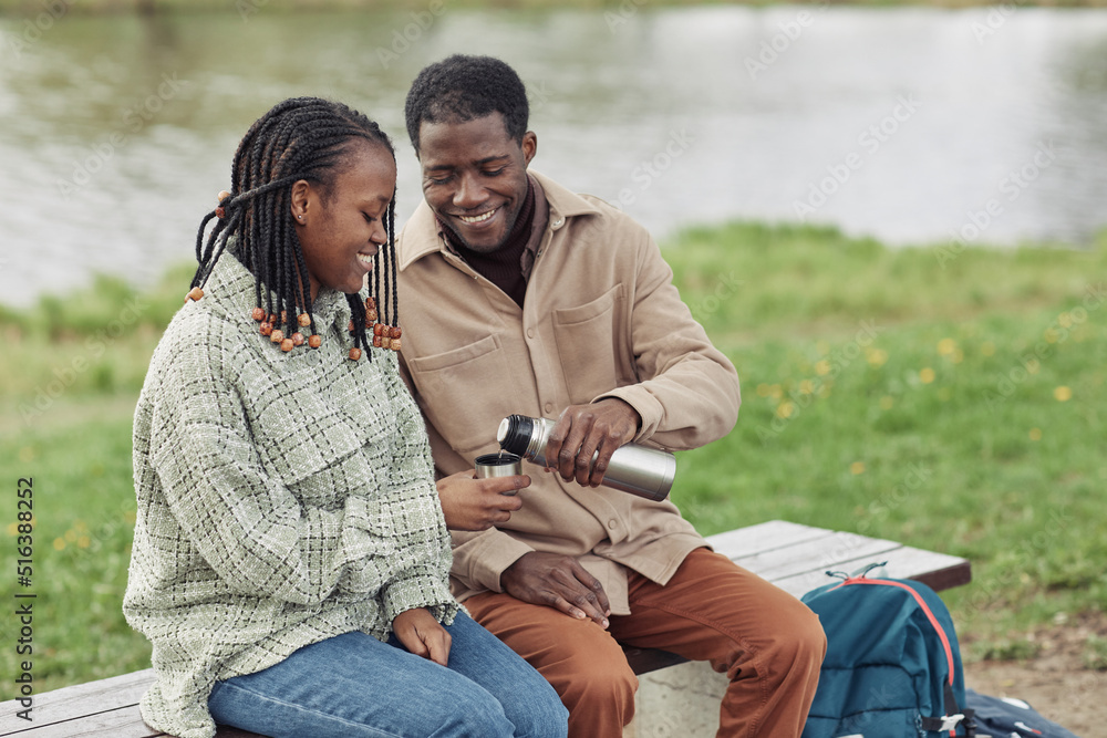 Happy African young couple sitting on bench and drinking hot tea from flask during their picnic in the park Happy African young couple sitting on bench and drinking hot tea from flask during their picnic in the park