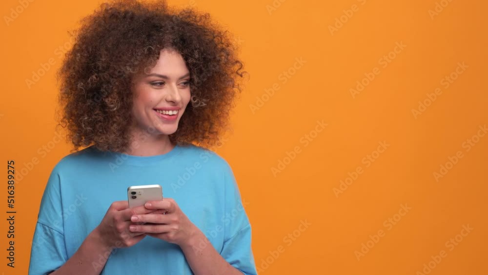 Cheerful curly haired blonde woman wearing blue t-shirt typing by phone in the orange studio Cheerful curly haired blonde woman wearing blue t-shirt typing by phone in the orange studio