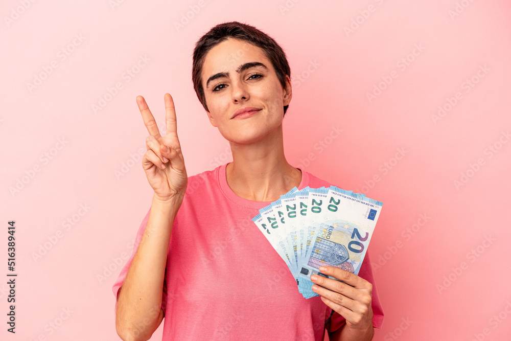 Young caucasian woman holding banknotes isolated on pink background showing number two with fingers. Young caucasian woman holding banknotes isolated on pink background showing number two with fingers.
