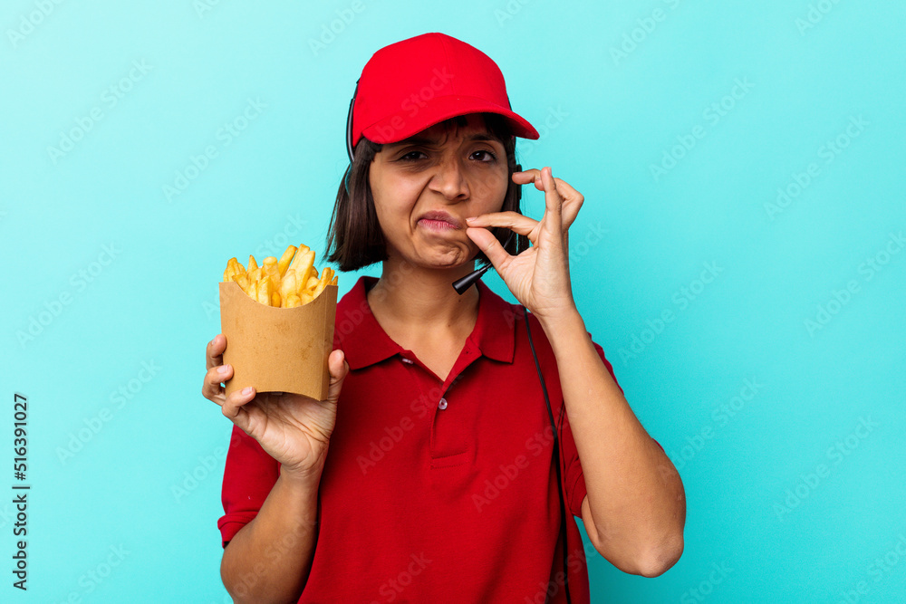 Young mixed race woman fast food restaurant worker holding fries isolated on blue background with fingers on lips keeping a secret. Young mixed race woman fast food restaurant worker holding fries isolated on blue background with fingers on lips keeping a secret.