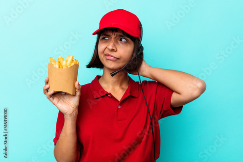 Young mixed race woman fast food restaurant worker holding fries isolated on blue background touching back of head, thinking and making a choice.