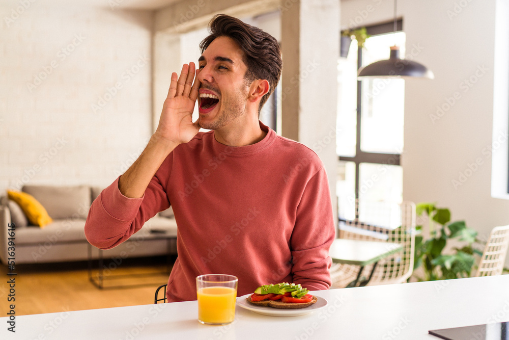 Young mixed race man having breakfast in his kitchen shouting and holding palm near opened mouth. Young mixed race man having breakfast in his kitchen shouting and holding palm near opened mouth.