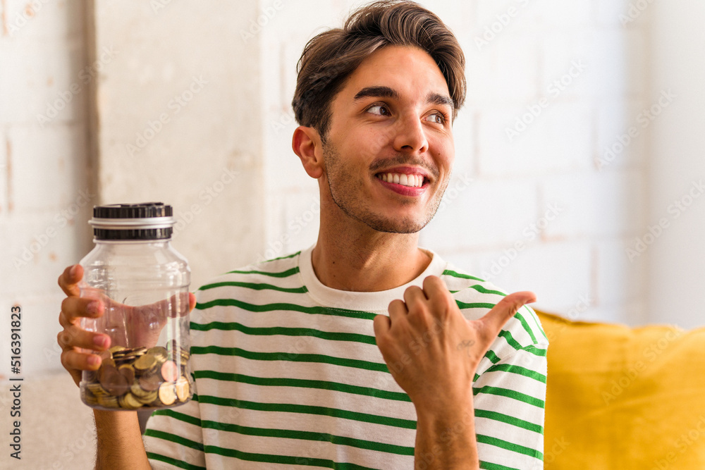 Young mixed race man holding piggy bank in his living room points with thumb finger away, laughing and carefree. Young mixed race man holding piggy bank in his living room points with thumb finger away, laughing and carefree.