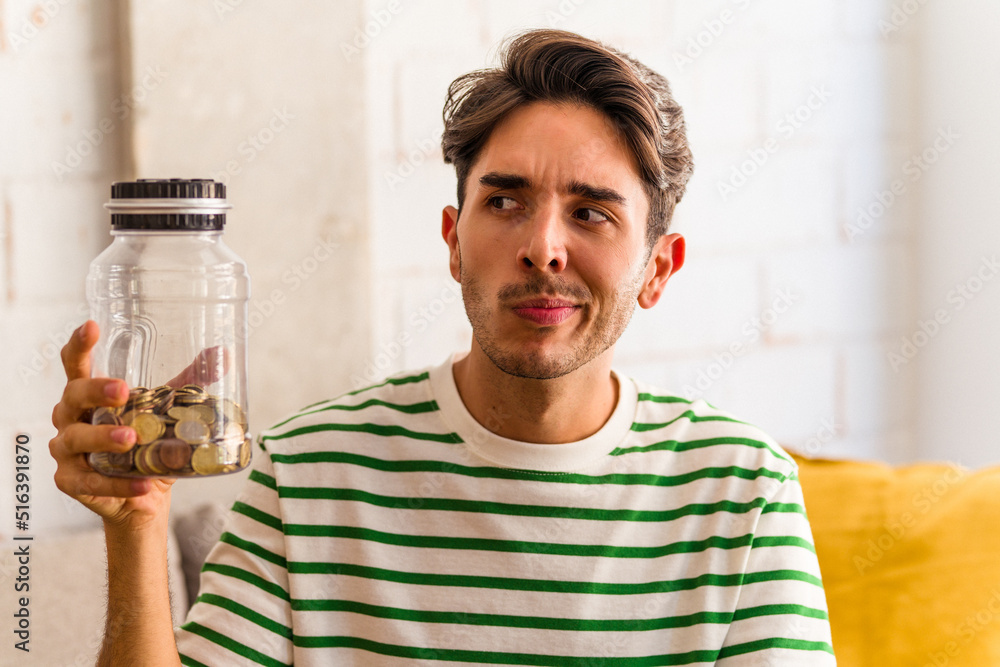 Young mixed race man holding piggy bank in his living room confused, feels doubtful and unsure. Young mixed race man holding piggy bank in his living room confused, feels doubtful and unsure.
