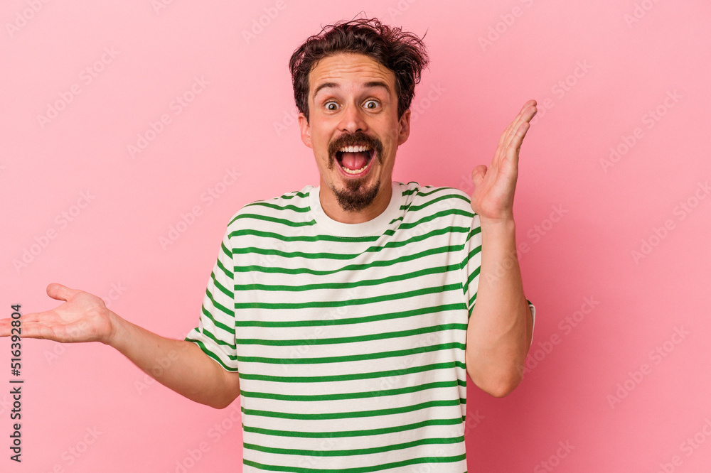 Young caucasian man isolated on pink background holds copy space on a palm, keep hand over cheek. Amazed and delighted.