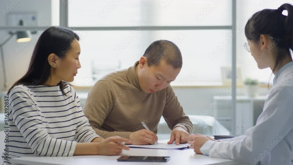Asian wife and husband signing medical agreement and talking with female doctor while visiting maternity clinic Asian wife and husband signing medical agreement and talking with female doctor while visiting maternity clinic