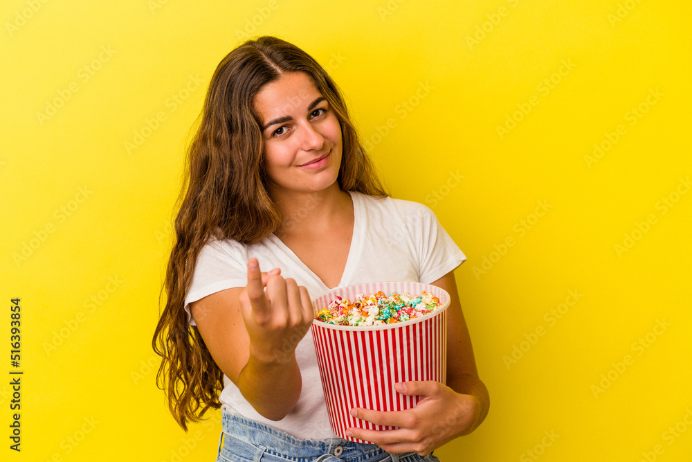 Young caucasian woman holding a popcorns isolated on yellow background  pointing with finger at you as if inviting come closer.