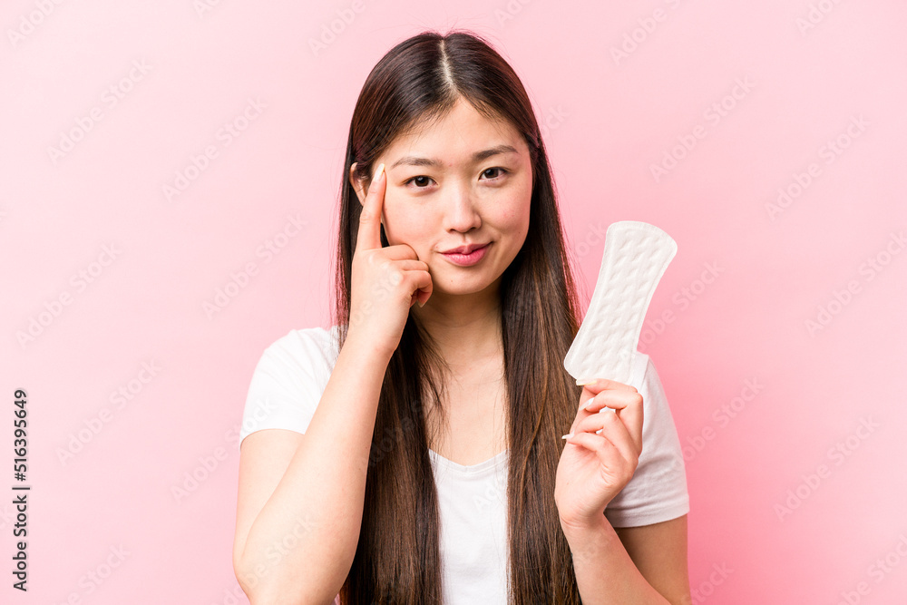 Young asian woman holding sanitary napkin isolated on pink background pointing temple with finger, thinking, focused on a task. Young asian woman holding sanitary napkin isolated on pink background pointing temple with finger, thinking, focused on a task.