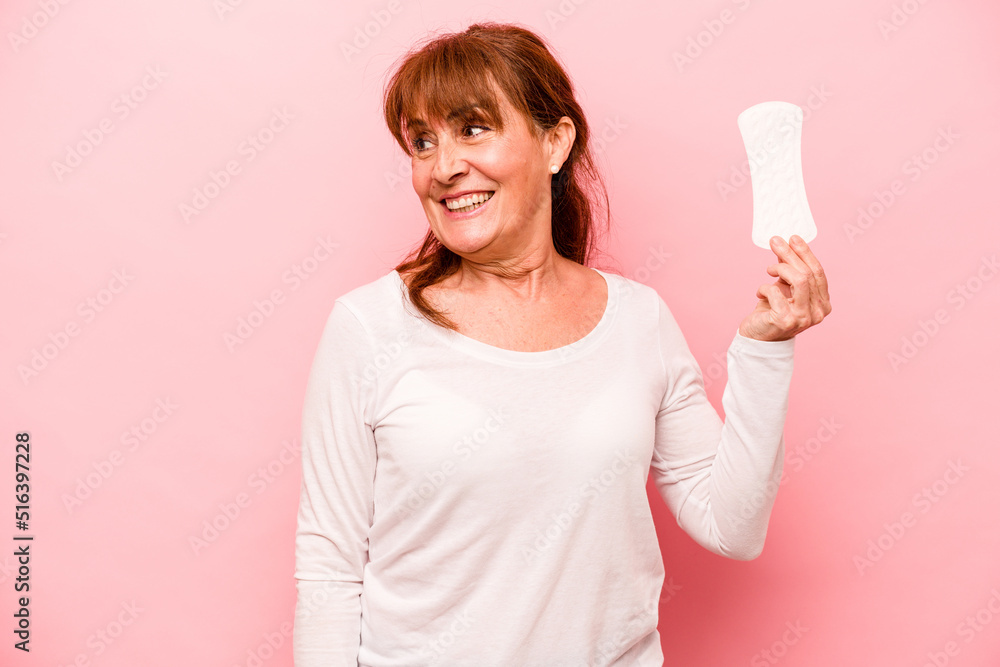 Middle age caucasian woman holding sanitary napkin isolated on pink background looks aside smiling, cheerful and pleasant. Middle age caucasian woman holding sanitary napkin isolated on pink background looks aside smiling, cheerful and pleasant.