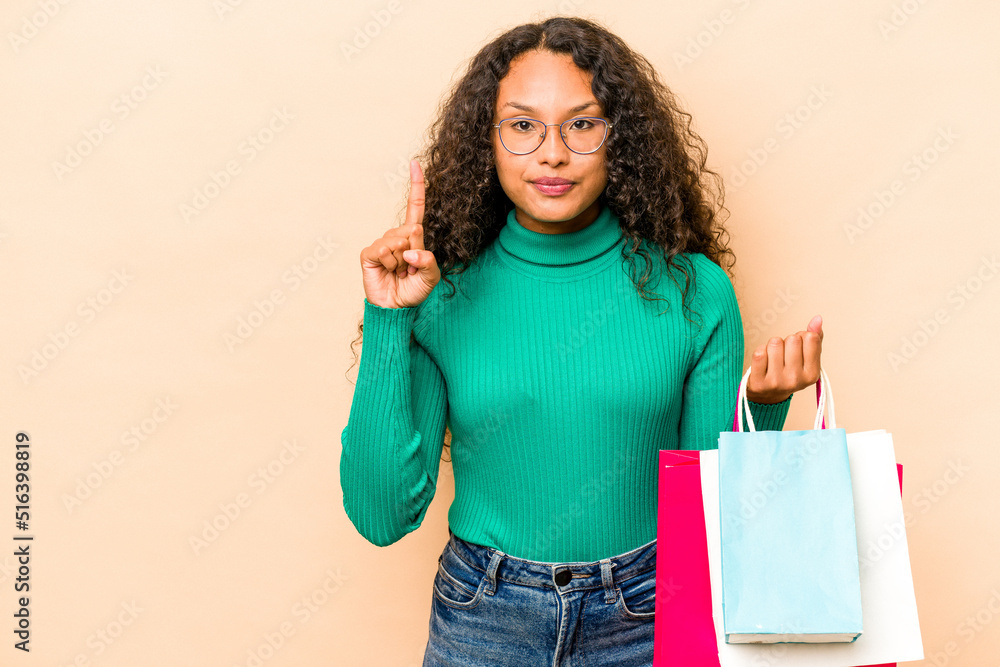 Young hispanic woman shopping some clothes isolated on beige background showing number one with finger. Young hispanic woman shopping some clothes isolated on beige background showing number one with finger.