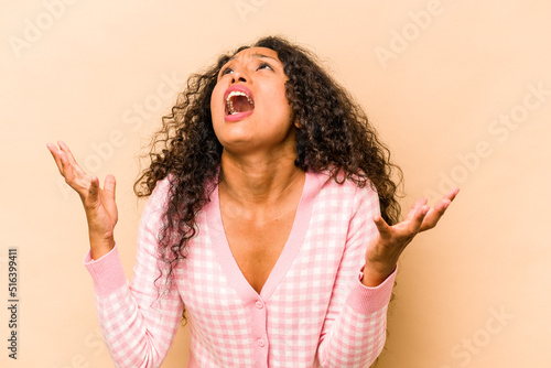 Young hispanic woman isolated on beige background screaming to the sky, looking up, frustrated.