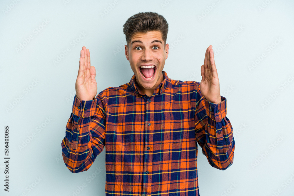 Naklejka premium Young caucasian man isolated on blue background receiving a pleasant surprise, excited and raising hands.