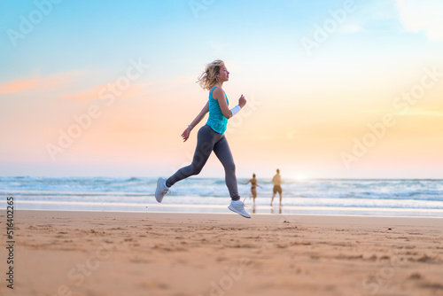 Active sporty woman run along ocean surf by water pool to keep fit and health. Sunset sand beach background with sun. Woman fitness, jogging workout and sport activity