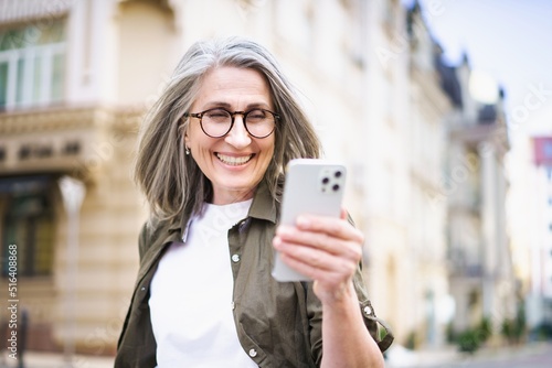 Beautiful mature woman with grey hair looking at white smartphone walking at the streets of old european town. Mature woman read text message or answering video call standing or walking outdoors
