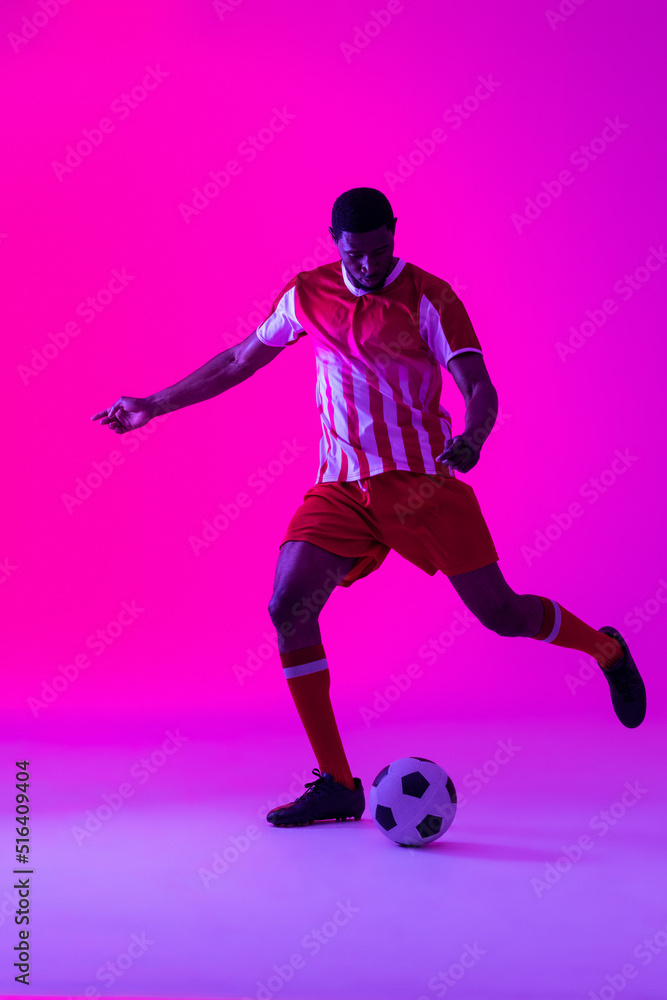 African american male soccer player kicking football over neon pink lighting