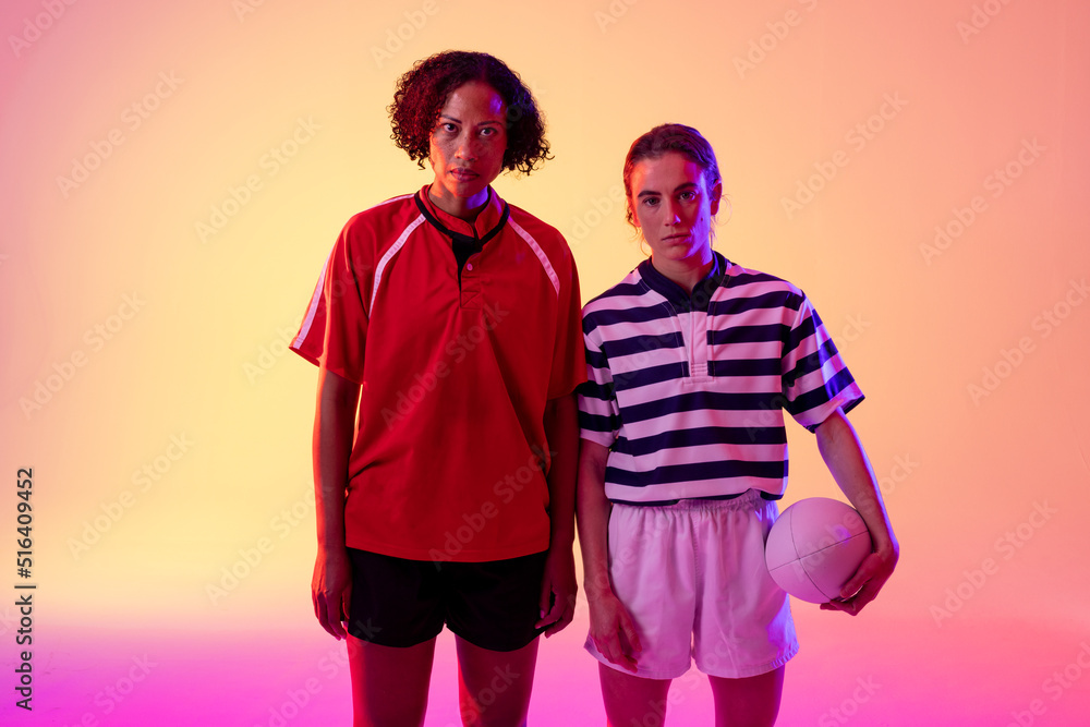 Obraz premium Portrait of two diverse female rugby players with rugby ball over neon pink lighting