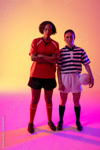 Portrait of two diverse female rugby players with rugby ball over neon pink lighting