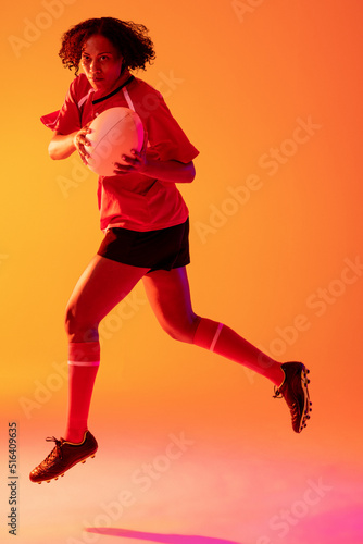 African american female rugby player with rugby ball over neon pink lighting
