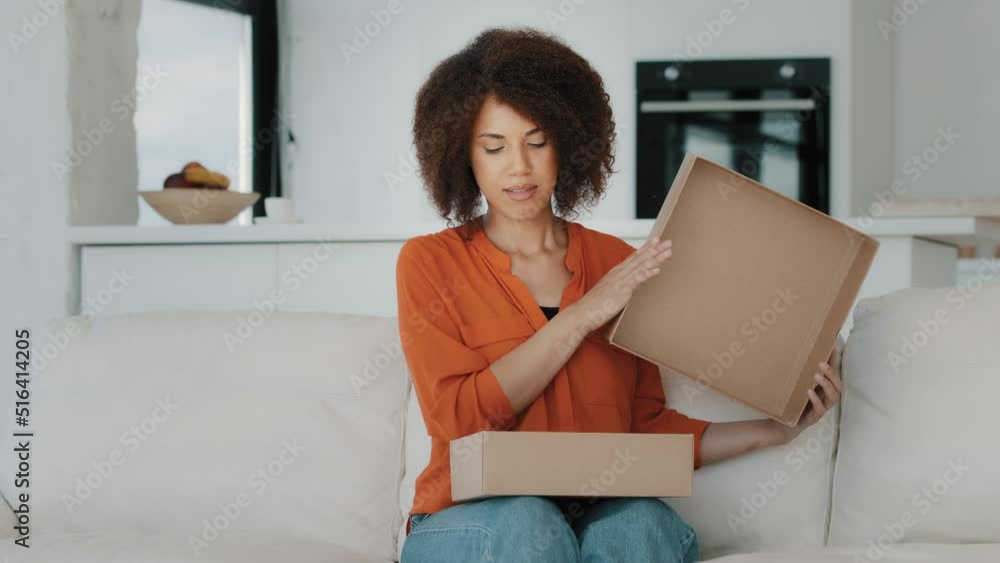 Curious African American buyer customer woman with curly hair sitting at couch at home unbox opening unpacking parcel box gift present purchase order from online shop shipping delivery service indoors Curious African American buyer customer woman with curly hair sitting at couch at home unbox opening unpacking parcel box gift present purchase order from online shop shipping delivery service indoors