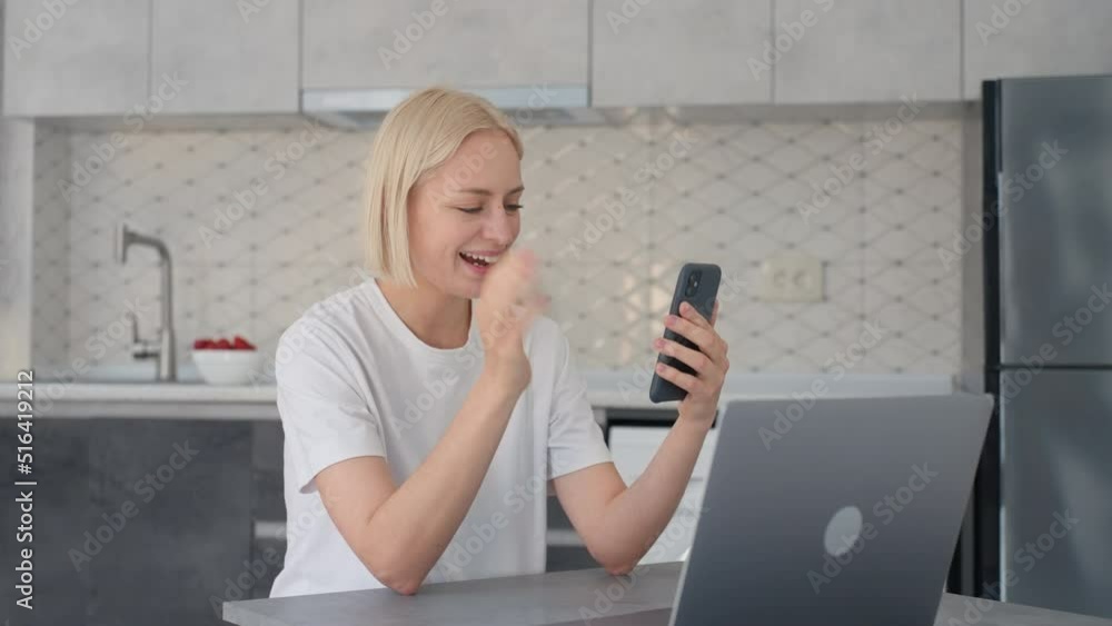 Joyful blonde woman waves bye in video call when sitting at laptop in kitchen Joyful blonde woman waves bye in video call when sitting at laptop in kitchen