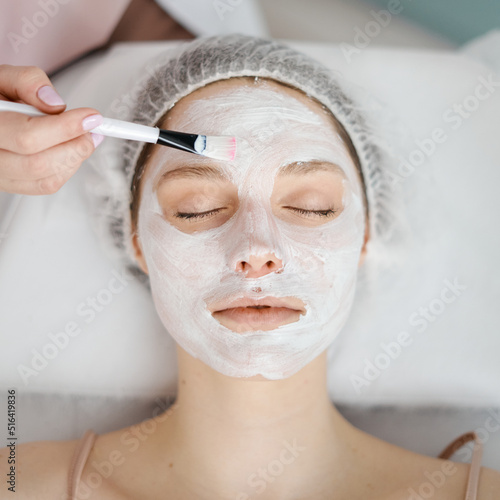 The young woman on a visit at the professional cosmetologist on a procedure for care of face skin.Cosmetician applying facial mask to the face of young beautiful woman in spa salon.