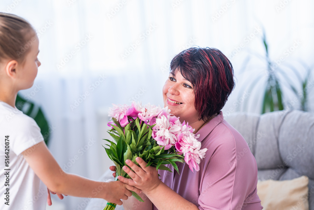 affectionate daughter gives bouquet of peonies to happy mother, congratulating her on Mother's Day during celebration of the holiday at home. loving child makes a pleasant surprise for mom on March 8.