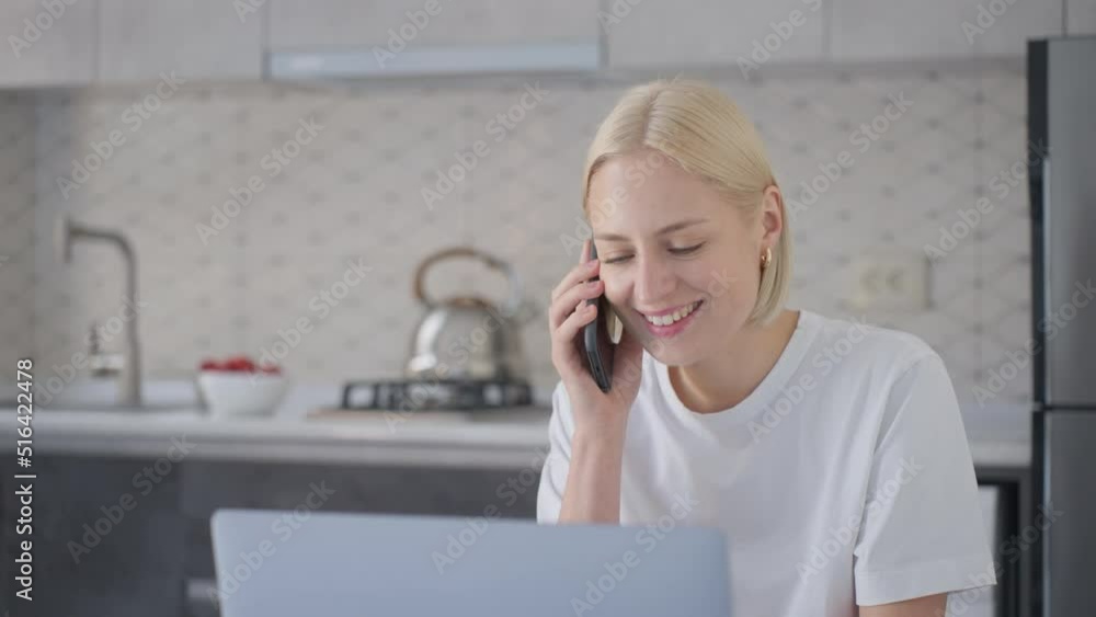 Young blonde woman starts phone talk when sitting at laptop in kitchen Young blonde woman starts phone talk when sitting at laptop in kitchen