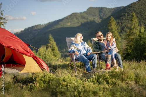 Young caucasian couple with little girl sit relaxed on chairs while traveling with tent in the mountains. Concept of happy family vacation on nature