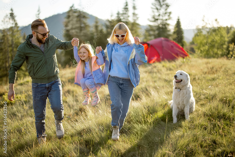 Fototapeta premium Young caucasian couple with little girl and dog have fun spending leisure time together while travel with tent in the mountains. Happy family summer vacation at campsite